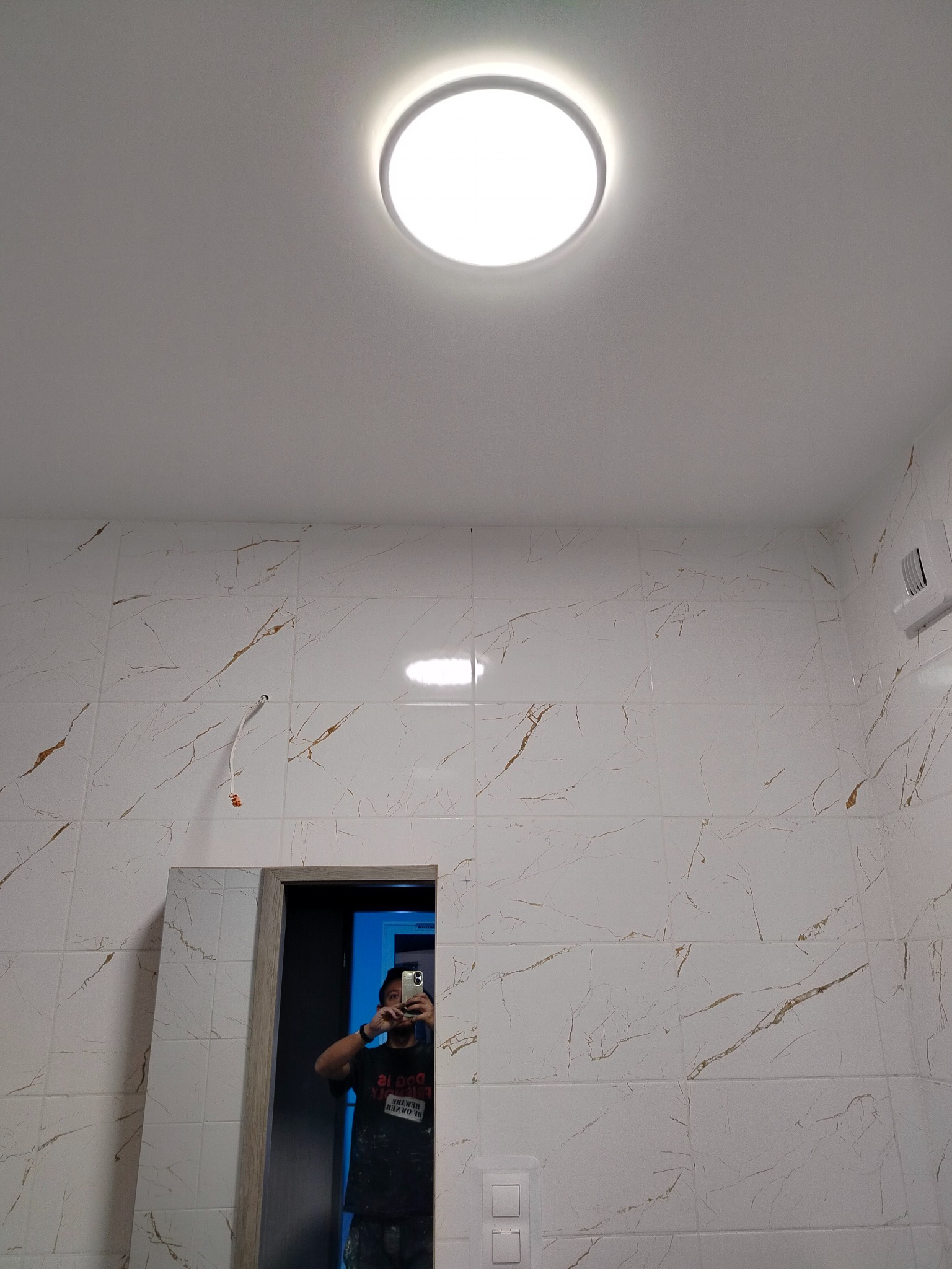 Wykończona łazienka z białymi płytkami imitującymi marmur, oświetlona okrągłą lampą sufitową. Widoczne lustro i instalacja. Osoba robi zdjęcie w lustrze.