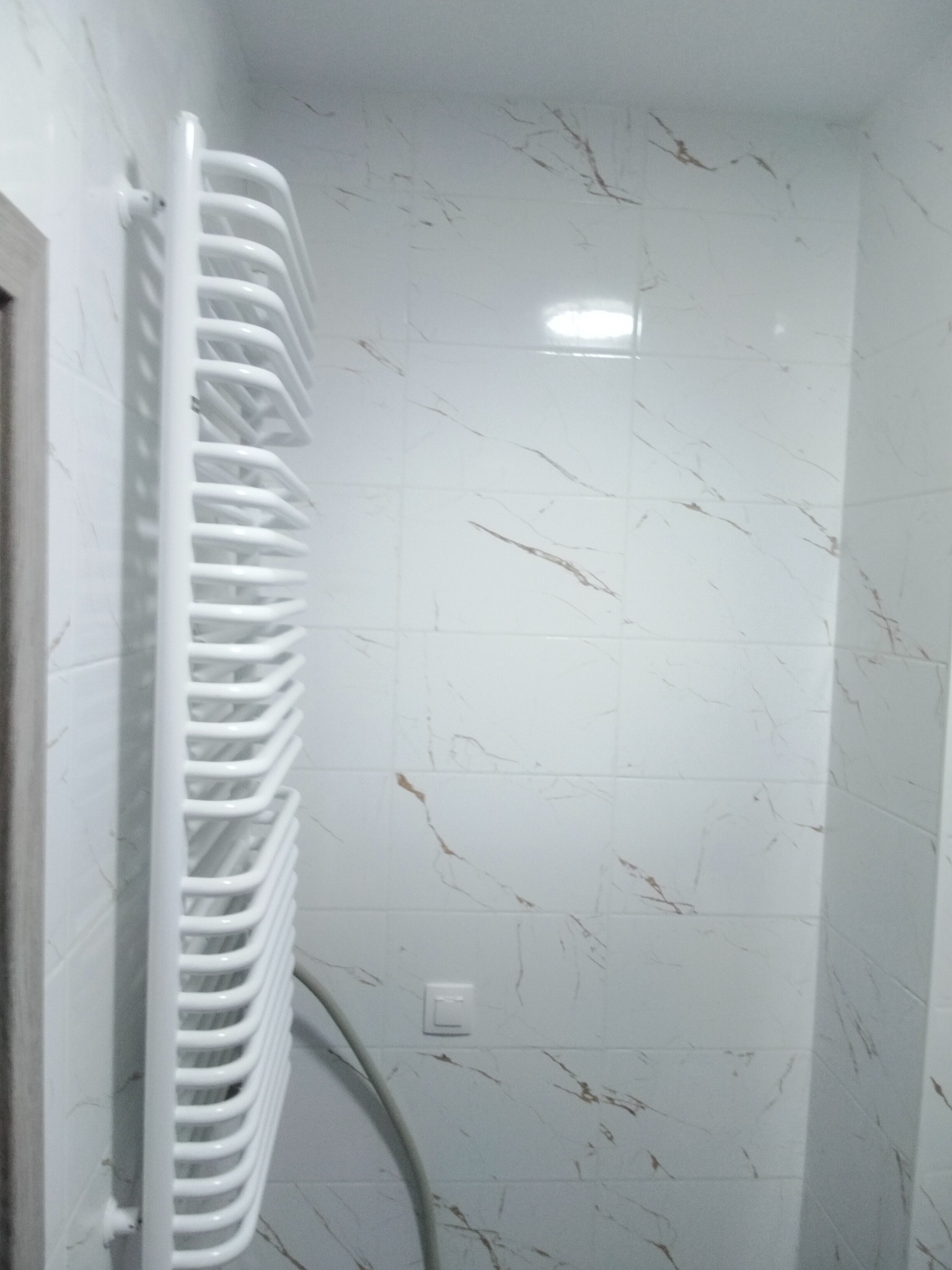 Biały grzejnik drabinkowy na ścianie z marmurowymi płytkami w łazience. Widoczny fragment drzwi i włącznik światła. Minimalistyczny design.