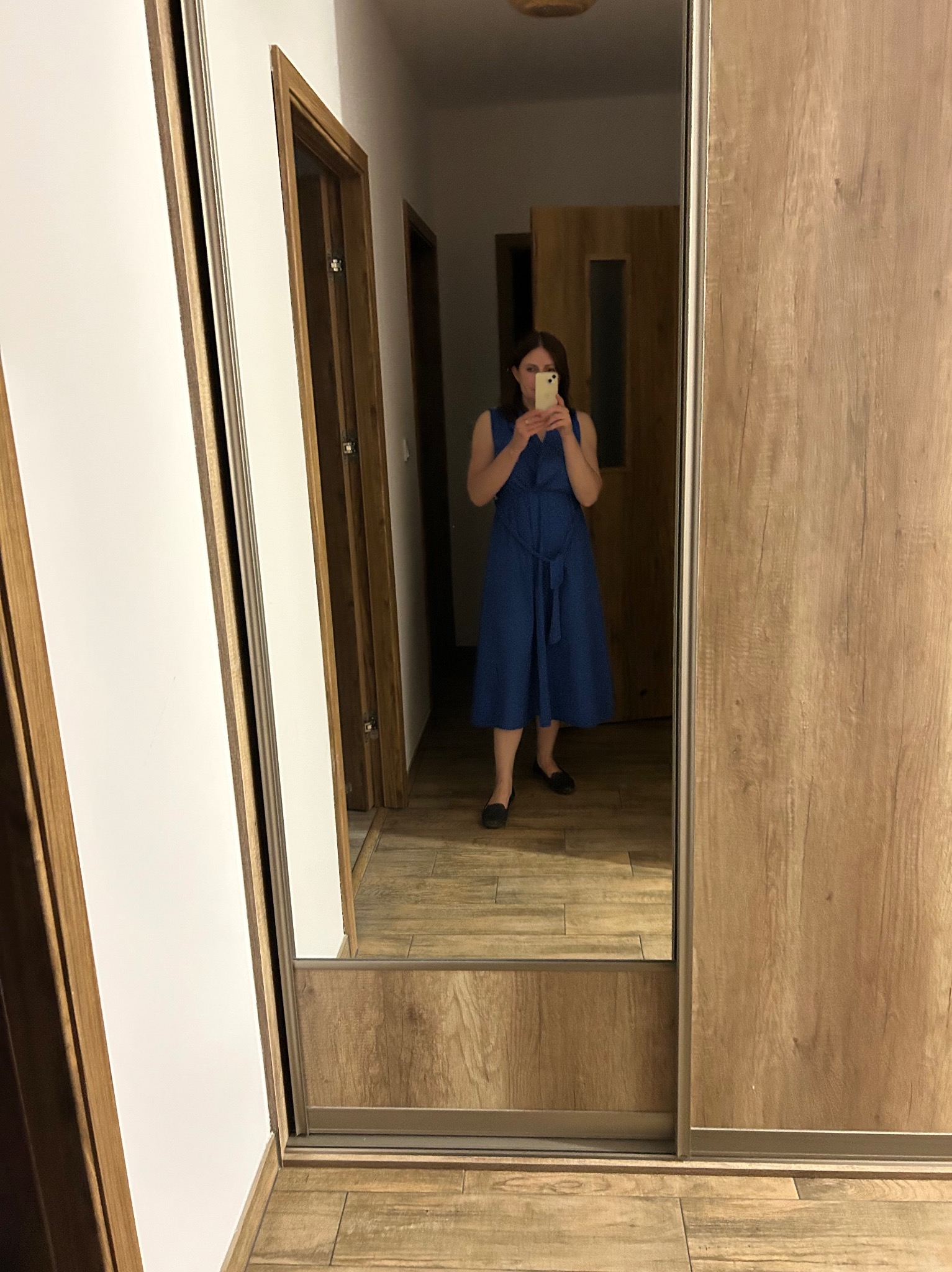 Odbicie kobiety w niebieskiej sukience w lustrze szafy przesuwnej z drewnianymi frontami. Widoczne drzwi i podłoga z płytek imitujących drewno.