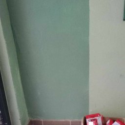 Antoni Pieńkos - Zamontowane, czerwone czujniki przeciwpożarowe na zielonej ścianie klatki schodowej, obok karton z nowymi urządzeniami. Widok z dołu, blisko drzwi wejściowych.