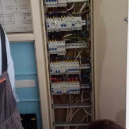 Antoni Pieńkos - Otwarta szafa elektryczna z widocznymi zabezpieczeniami, przewodami i elementami instalacji. Fragment muru z przewodami w tle. Widoczny fragment osoby w ubraniu roboczym.