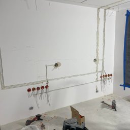 Białe ściany z rozrysowaną instalacją elektryczną, widoczne kable, puszki i narzędzia na podłodze. Etap prac przed wykończeniem.