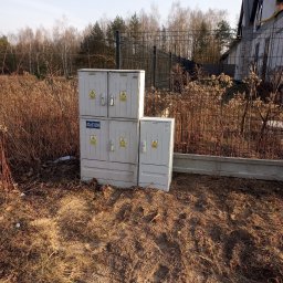 Antoni Pieńkos - Zewnętrzna szafa elektryczna z oznaczeniami ostrzegawczymi, umieszczona na gruncie w pobliżu ogrodzenia i budynków w stanie surowym. Widoczne krzaki i drzewa w tle.