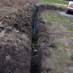 Antoni Pieńkos - Wykop w ziemi z niebieską rurą w środku, widoczny fragment koparki i narzędzia. Prace ziemne w terenie otwartym, w tle pole i drzewa.