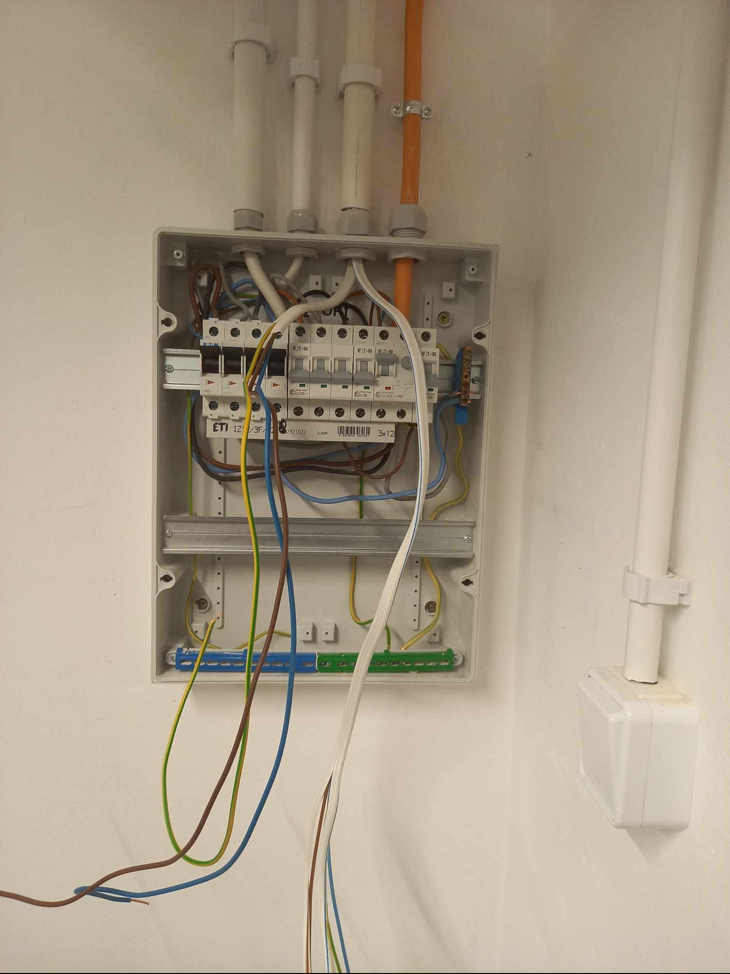 Otwarta skrzynka elektryczna z bezpiecznikami ETI na białej ścianie, widoczne kable w różnych kolorach i rury instalacyjne. Obok puszka elektryczna.