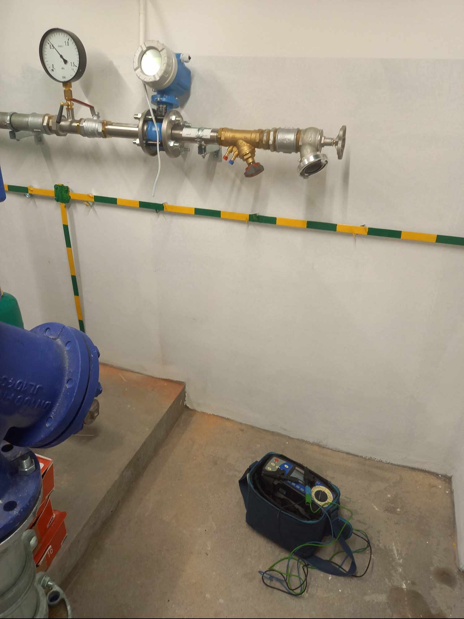 Pomiar instalacji wodnej: manometr, przepływomierz i rury z żółto-zielonym uziemieniem. Na podłodze leży miernik w torbie.