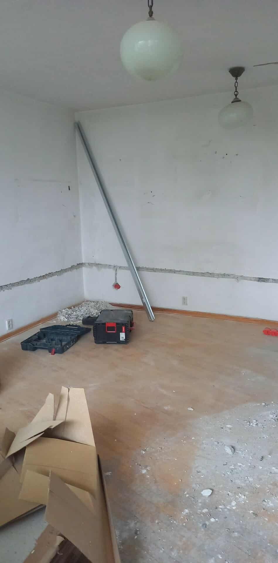 Wnętrze w trakcie remontu z widocznymi bruzdami w ścianach, puszkami elektrycznymi, narzędziami i profilem aluminiowym opartym o ścianę. Dwie lampy sufitowe.