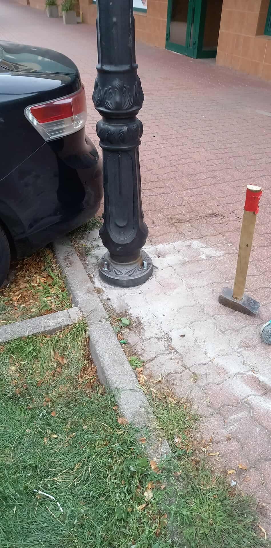 Montaż ozdobnej latarni ulicznej na chodniku z kostki, obok zaparkowanego auta. Widoczny młotek z drewnianym trzonkiem i czerwoną taśmą.