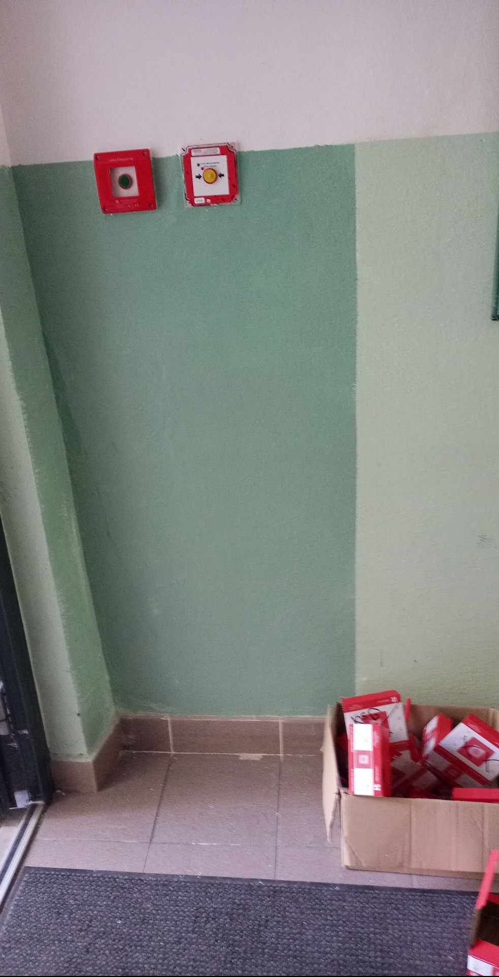 Zamontowane, czerwone czujniki przeciwpożarowe na zielonej ścianie klatki schodowej, obok karton z nowymi urządzeniami. Widok z dołu, blisko drzwi wejściowych.
