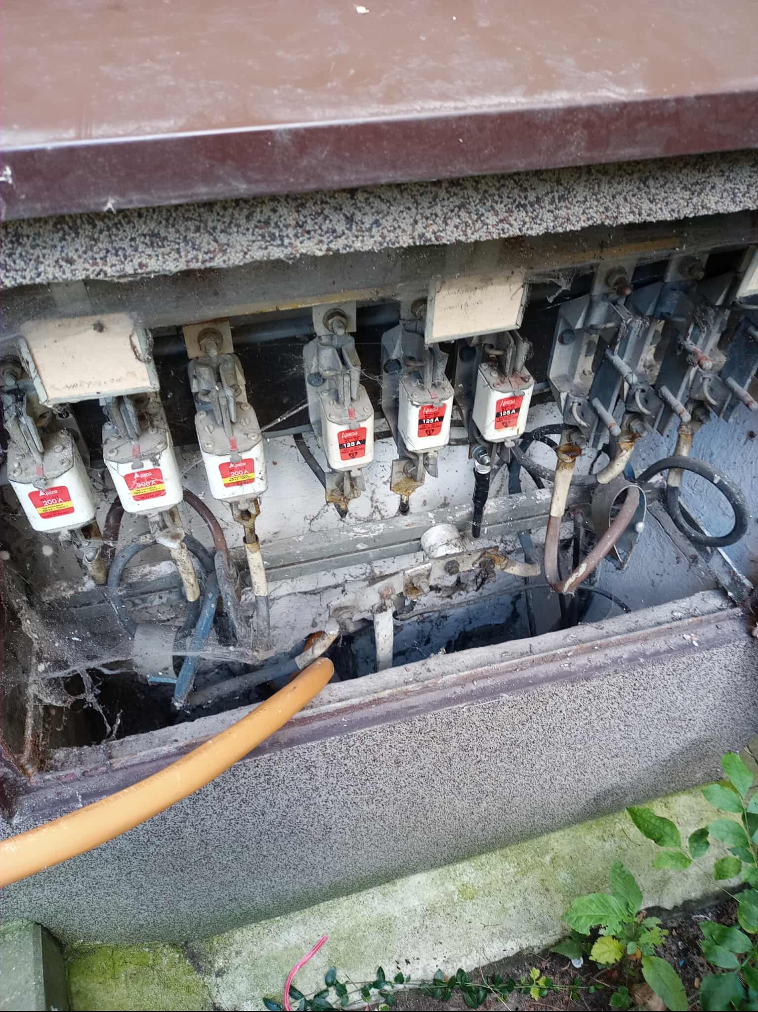 Otwarta skrzynka z bezpiecznikami, widoczne stare bezpieczniki 200A i 125A, okablowanie i rura instalacyjna. Widok z góry na instalację elektryczną w szarej obudowie.