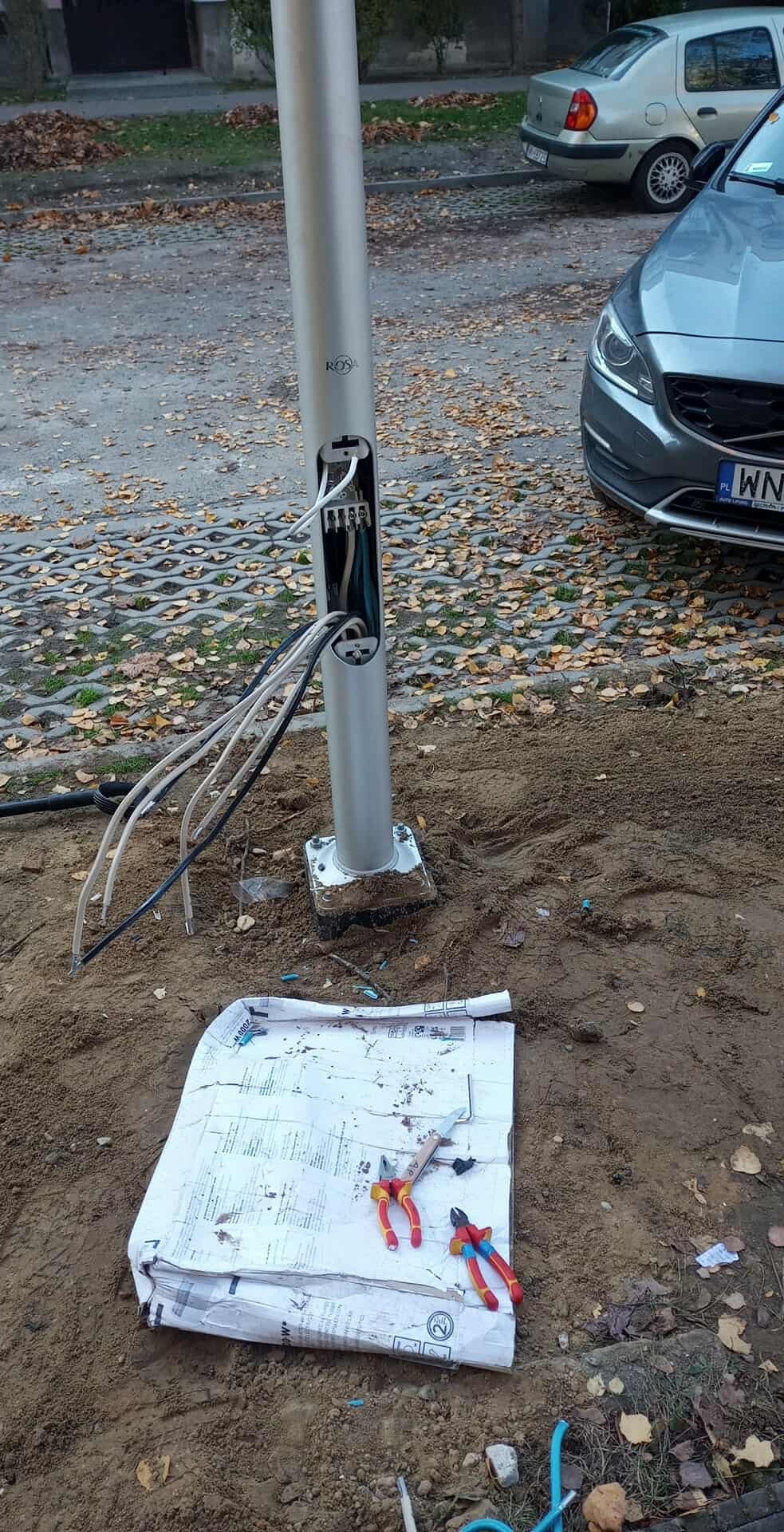 Srebrny słup oświetleniowy z otwartą skrzynką przyłączeniową, widoczne przewody. Na ziemi rozłożone plany i narzędzia, w tle zaparkowany samochód.