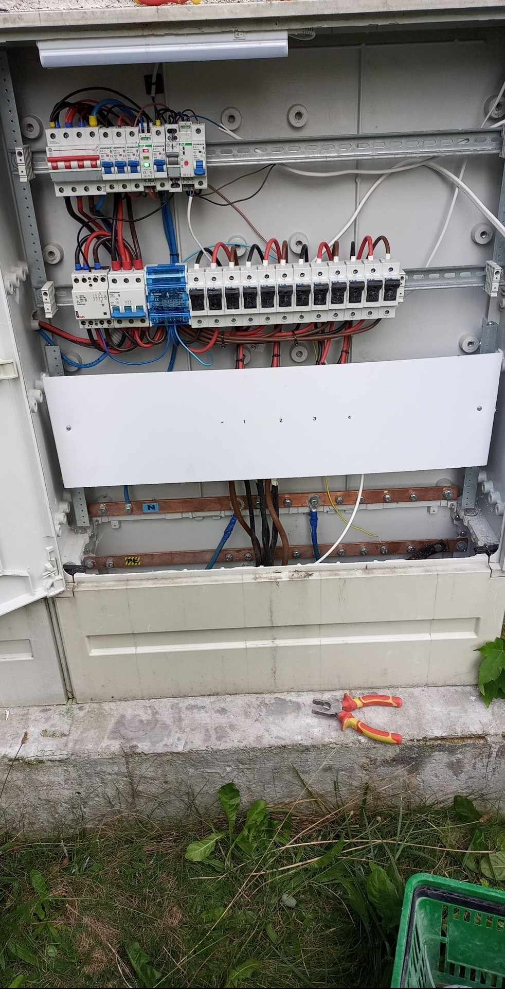 Otwarta skrzynka z zabezpieczeniami i przewodami elektrycznymi. Widoczne bezpieczniki, listwy zaciskowe i kable. Na dole leżą kombinerki z żółto-czerwoną rękojeścią.