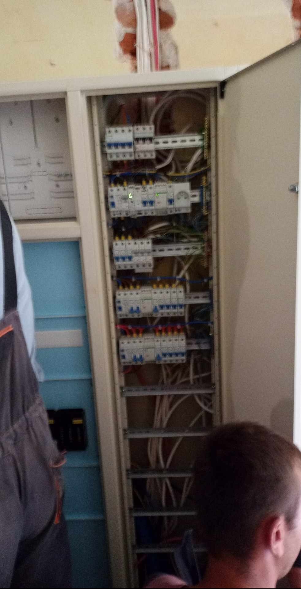 Otwarta szafa elektryczna z widocznymi zabezpieczeniami, przewodami i elementami instalacji. Fragment muru z przewodami w tle. Widoczny fragment osoby w ubraniu roboczym.