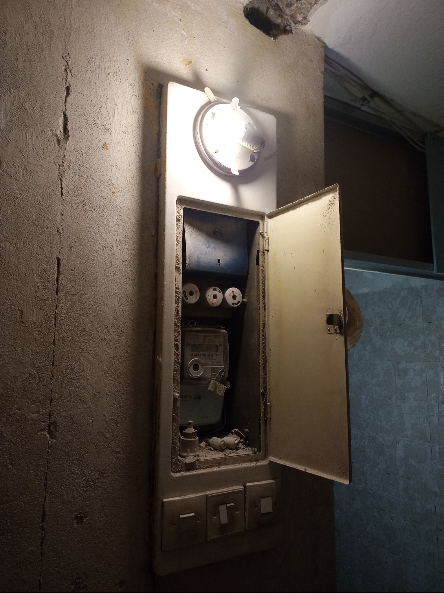 Otwarta skrzynka elektryczna z licznikiem i bezpiecznikami na starej, popękanej ścianie. Widoczne trzy włączniki. Oświetlenie wewnątrz skrzynki.