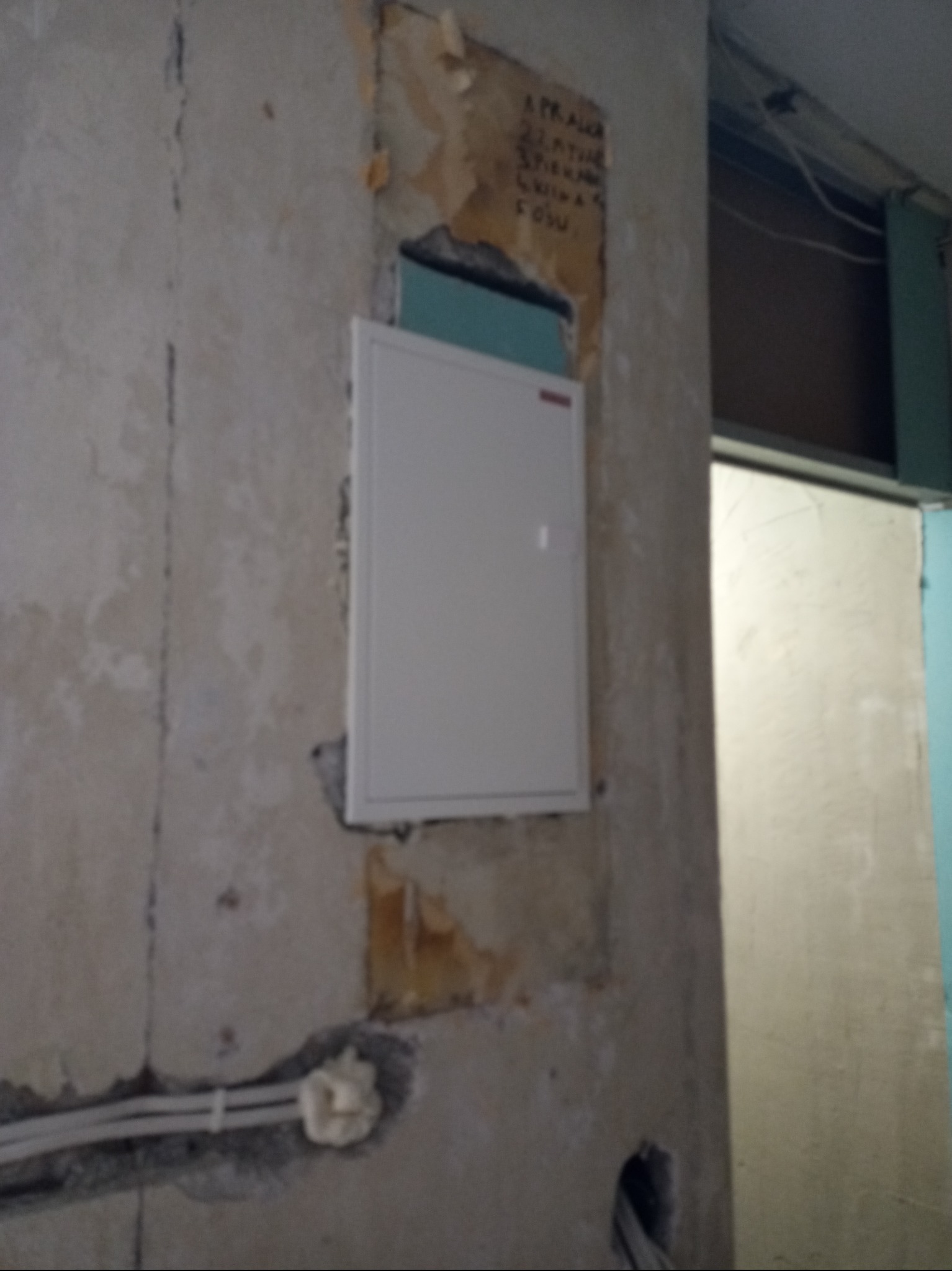 Biała skrzynka elektryczna zamontowana na surowej ścianie z widocznymi przewodami i resztkami tapety, obok framuga drzwi.