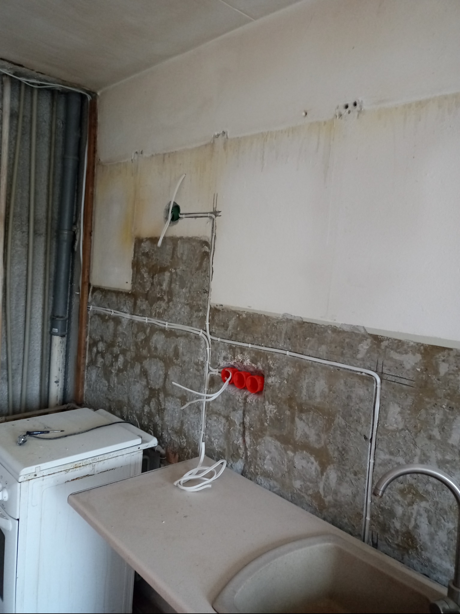 Ściana w trakcie remontu z widocznymi przewodami elektrycznymi, puszkami i fragmentami instalacji. Widoczny fragment pralki i zlewu.