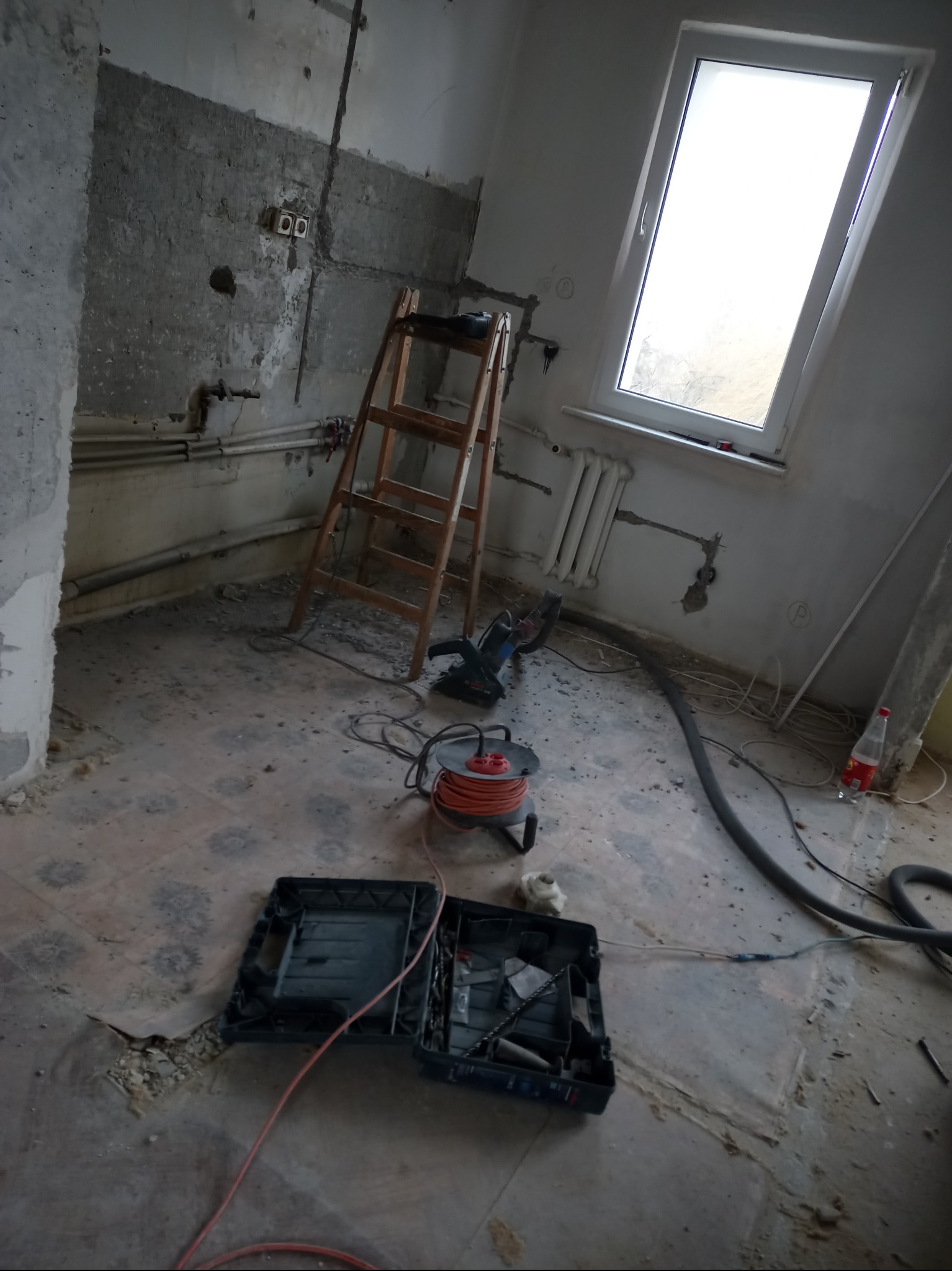 Remont pokoju: odsłonięte kable, puszki elektryczne, drabina, przedłużacz, walizka z narzędziami. Widoczne ślady po kuciu w ścianach i podłodze.