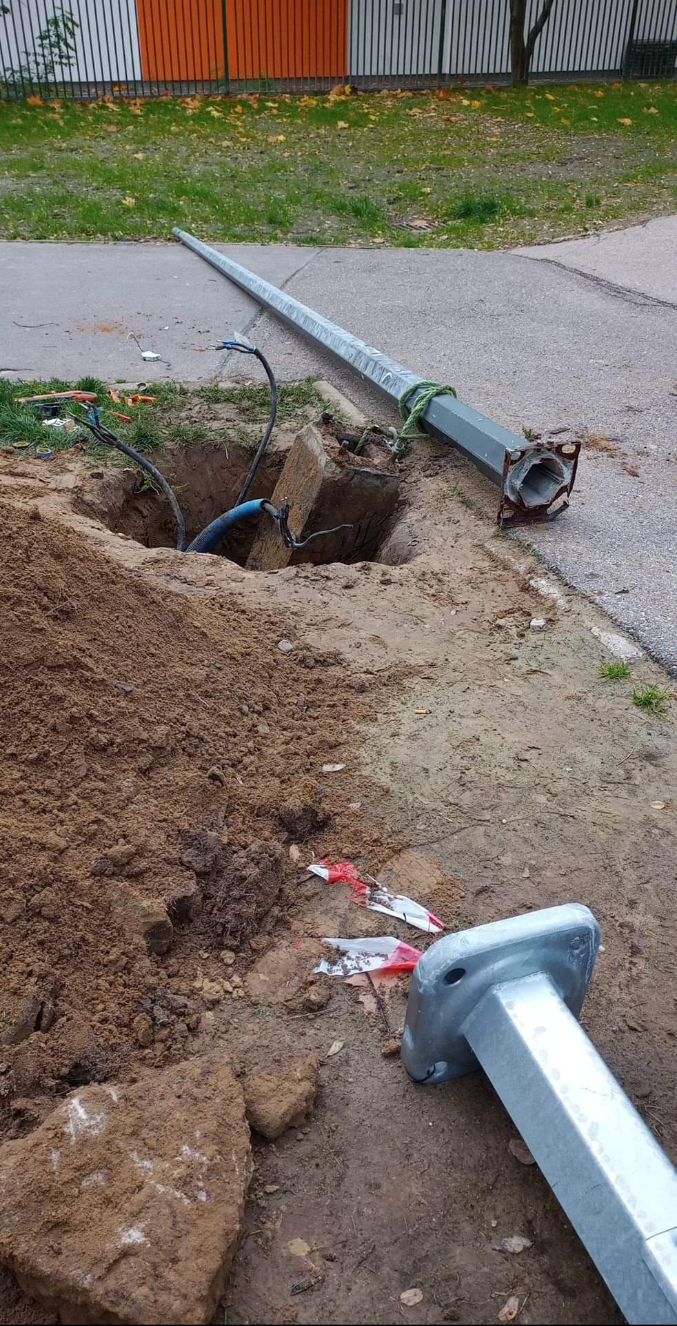 Uszkodzony słup oświetleniowy leżący obok wykopu z wystającymi kablami. Widoczne elementy demontażu i zabezpieczenia terenu.