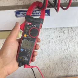 Miernik cęgowy UNI-T UT216C w dłoni, pomiar napięcia instalacji fotowoltaicznej. Wyświetlacz pokazuje 13.15. Widoczne kable i złącza instalacji.