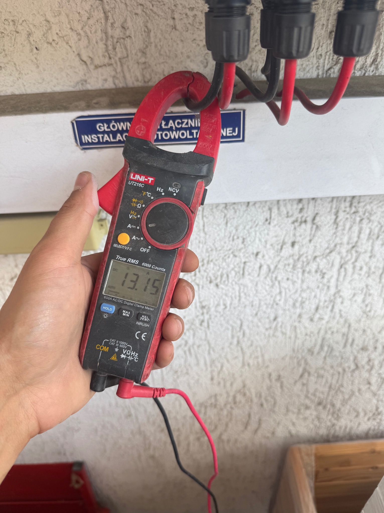 Miernik cęgowy UNI-T UT216C w dłoni, pomiar napięcia instalacji fotowoltaicznej. Wyświetlacz pokazuje 13.15. Widoczne kable i złącza instalacji.