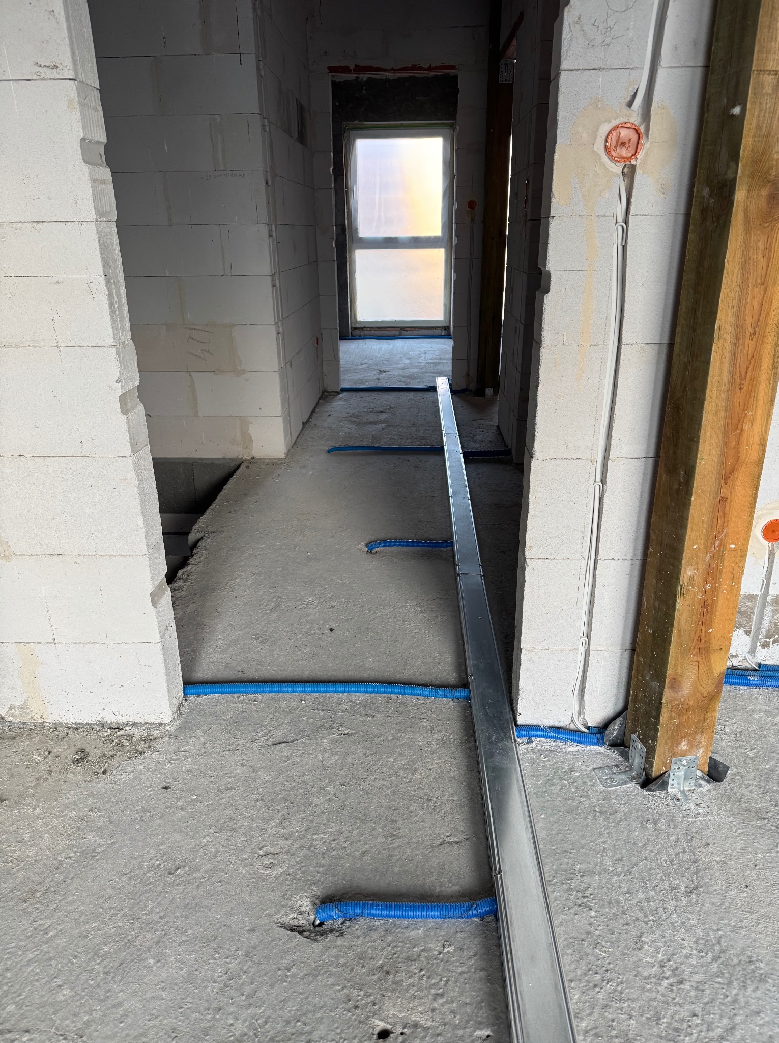 Niebieskie rury instalacyjne w betonowej posadzce, prowadzone wzdłuż metalowej szyny montażowej w surowym wnętrzu budynku w trakcie budowy. Widoczne puszki elektryczne.