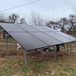 TWOJA SPÓŁKA SPÓŁKA Z OGRANICZONĄ ODPOWIEDZIALNOŚCIĄ - Montaż paneli fotowoltaicznych na metalowej konstrukcji w ogrodzie. Widoczne drabiny, narzędzia. Tło stanowią drzewa i budynek z czerwonej cegły.
