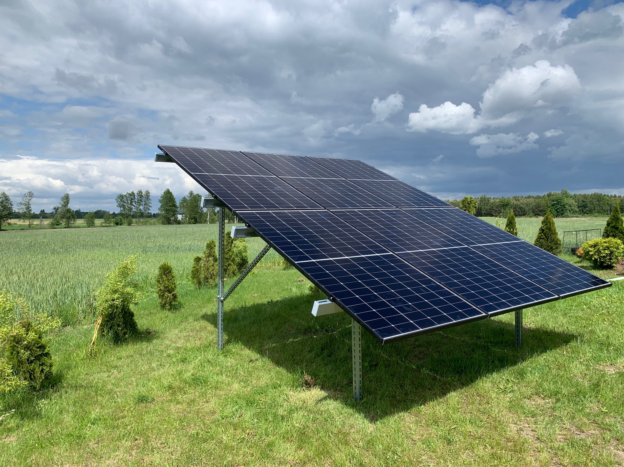 Instalacja fotowoltaiczna na gruncie, widok na panele w krajobrazie rolnym pod pochmurnym niebem. Widoczne metalowe wsporniki i zielone otoczenie.