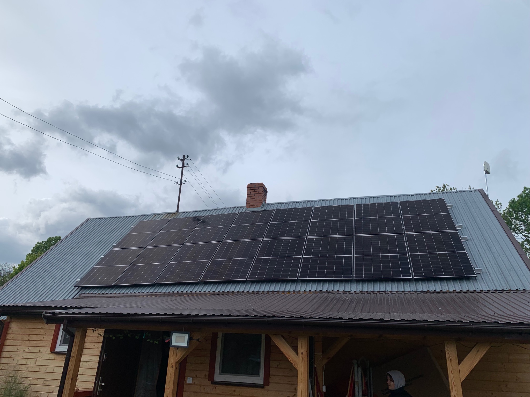 Panele słoneczne na blaszanym dachu drewnianego domu z ceglanym kominem, pochmurne niebo w tle. Widoczny słup energetyczny.