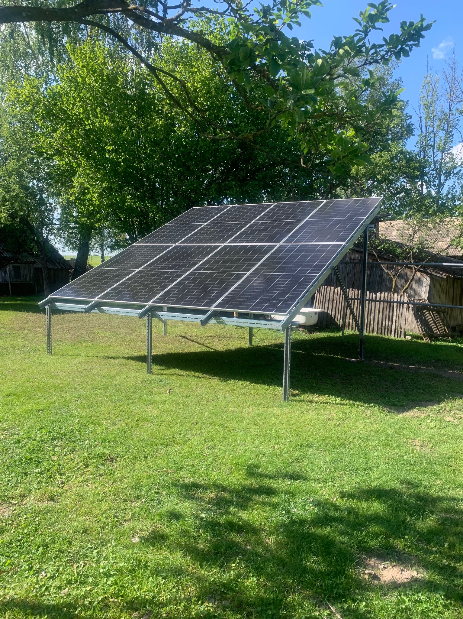 Panele fotowoltaiczne na metalowej konstrukcji, zainstalowane na trawniku w wiejskim otoczeniu, z widocznymi drzewami i budynkiem gospodarczym w tle.