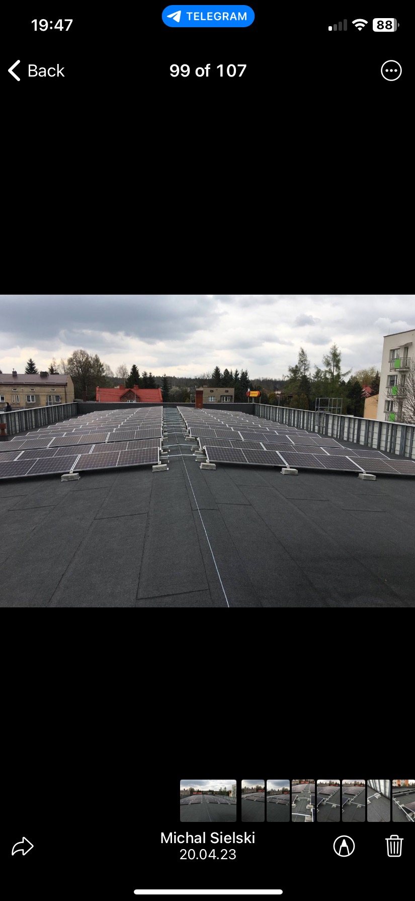 Widok z góry na dużą instalację fotowoltaiczną na płaskim dachu budynku. Panele ułożone w rzędach, widoczne elementy konstrukcyjne i tło zabudowy miejskiej.