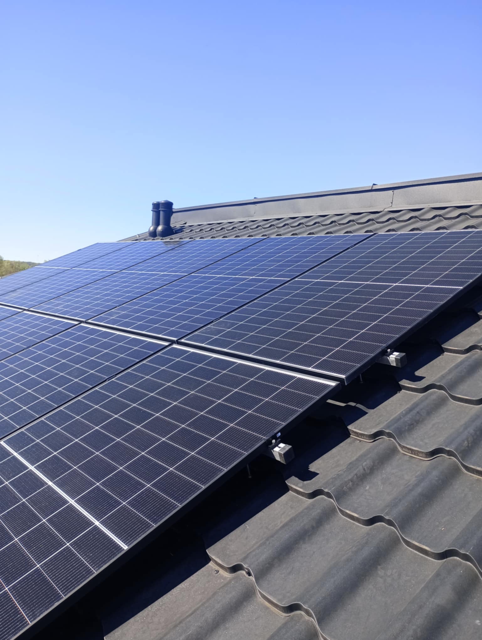 Panele fotowoltaiczne na dachu z ciemnej dachówki ceramicznej w słoneczny dzień. Widoczny komin. Układ paneli w orientacji poziomej.