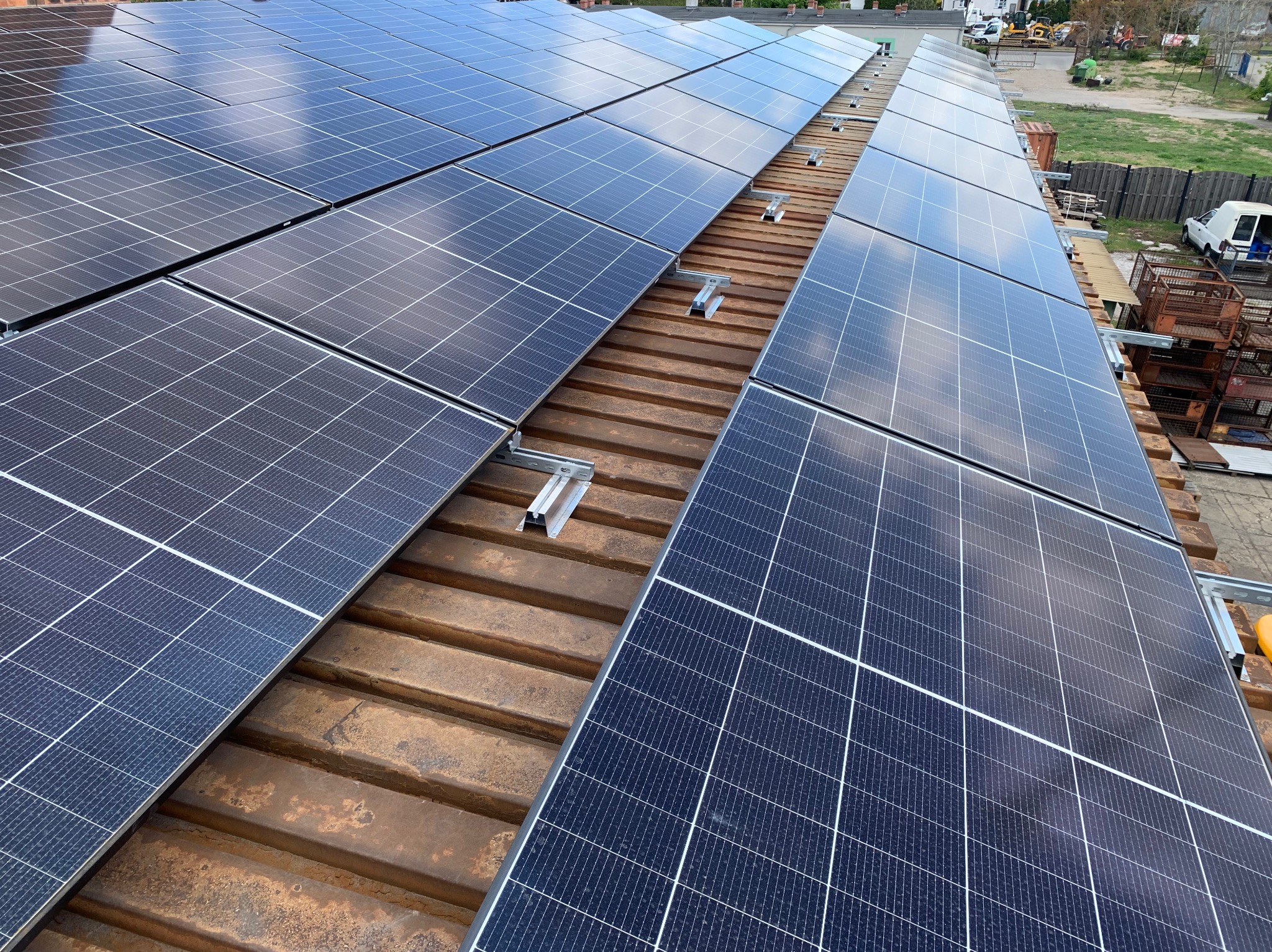 Panele fotowoltaiczne na brązowym, blaszanym dachu z widocznymi mocowaniami i odbiciem nieba, z fragmentami otoczenia w tle. Ujęcie z góry.