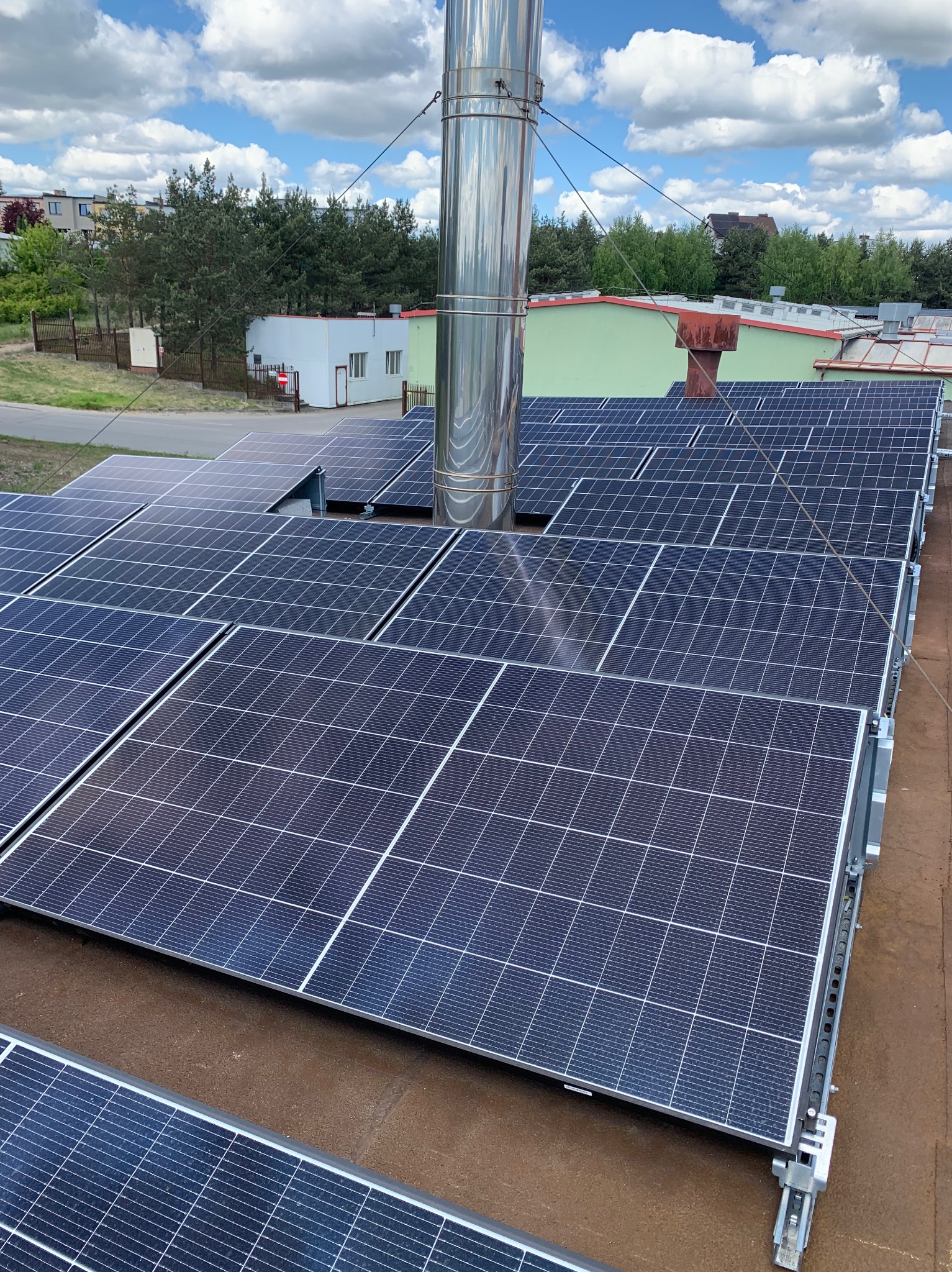 Instalacja fotowoltaiczna na dachu budynku z kominem, widok z góry. Panele w rzędach, wyraźne niebieskie krzemowe ogniwa, mocowania. Słoneczny dzień, błękitne niebo z chmurami.