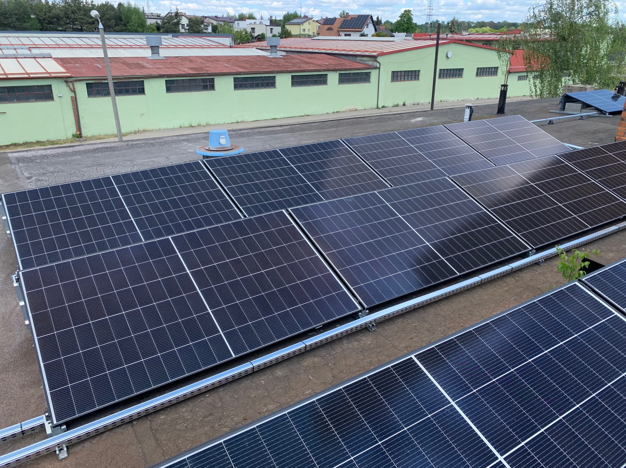 Panele fotowoltaiczne na dachu budynku przemysłowego. Widoczne elementy montażowe i tło z zielonym budynkiem oraz drzewami. Ujęcie z góry.