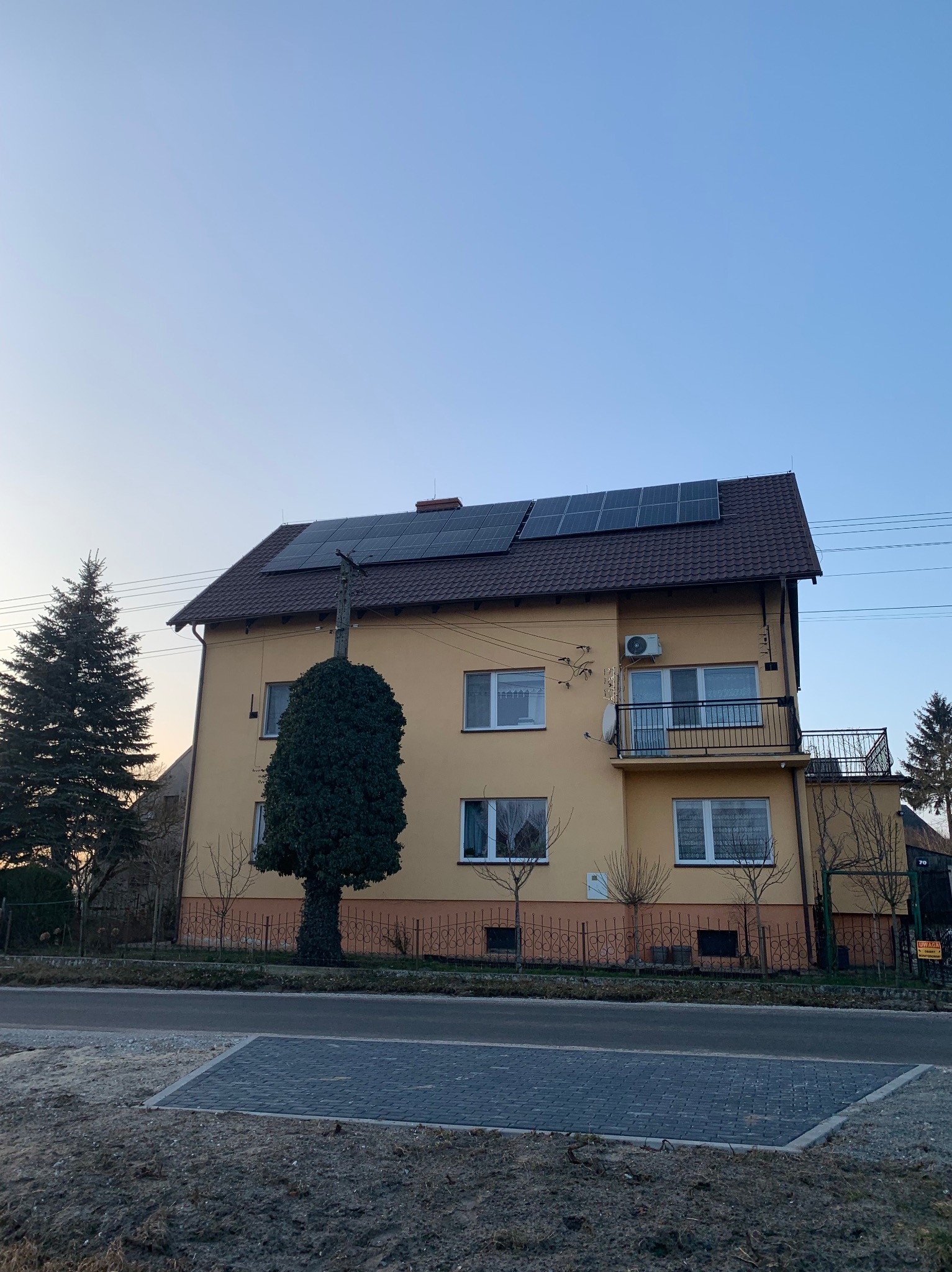 Żółty dom z brązowym dachem i panelami słonecznymi w Warszawie. Na balkonie klimatyzator. Uwagę przykuwa duży, zielony krzew przed budynkiem.