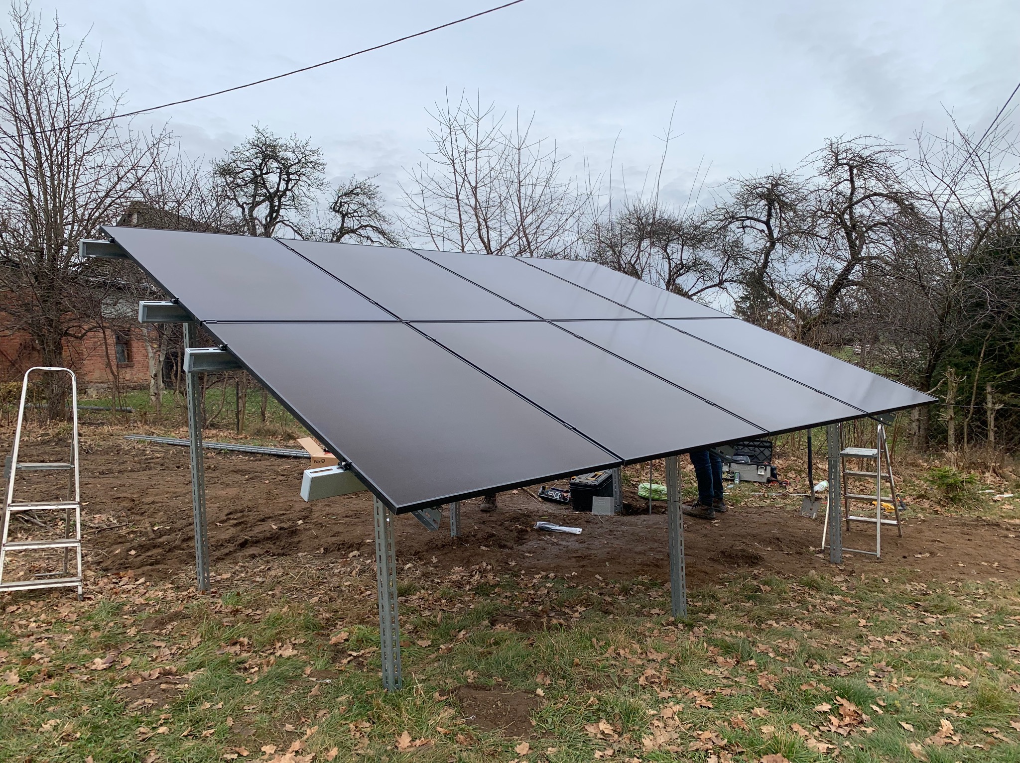 Montaż paneli fotowoltaicznych na metalowej konstrukcji w ogrodzie. Widoczne drabiny, narzędzia. Tło stanowią drzewa i budynek z czerwonej cegły.