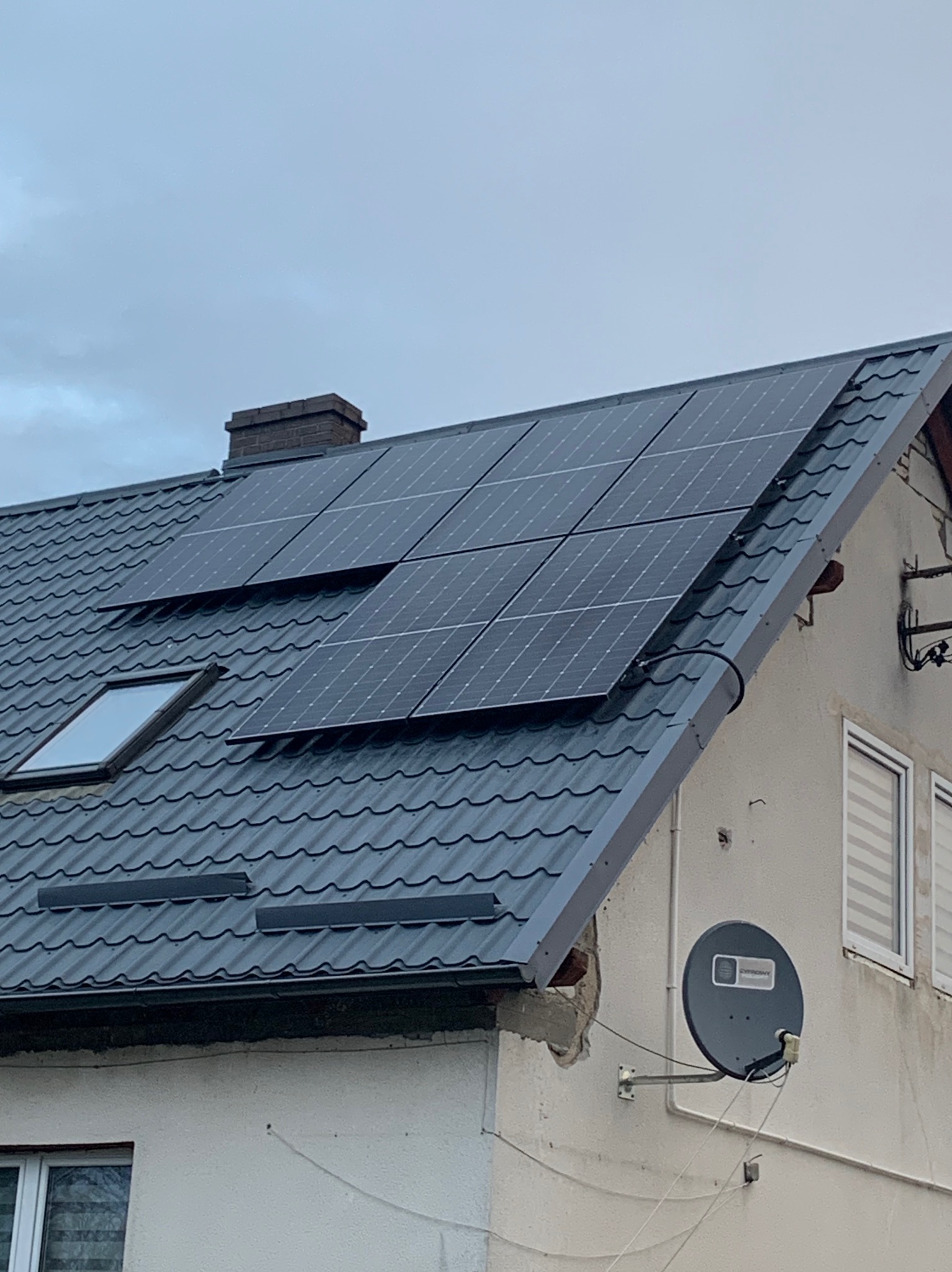 Panele słoneczne na dachu domu z szarej dachówki, widoczne okno dachowe i komin. Na ścianie budynku antena satelitarna.