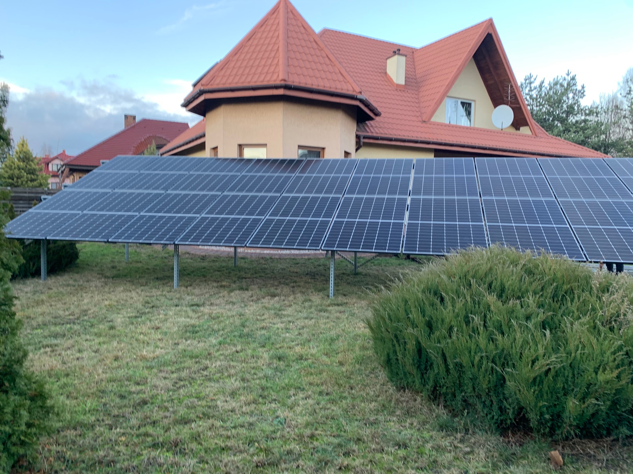Panele fotowoltaiczne na trawniku przed domem z czerwoną dachówką, widoczne krzewy i drzewa. Instalacja naziemna, w tle budynek mieszkalny.
