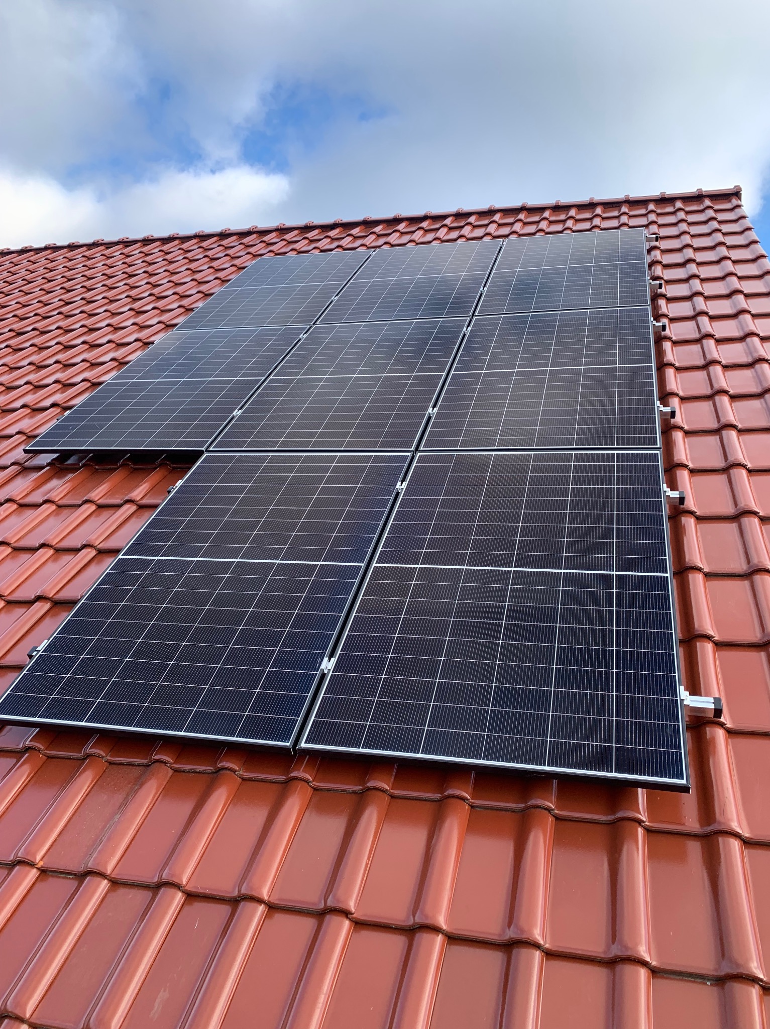 Panele słoneczne na dachu z czerwonej dachówki, widok z dołu pod kątem. Błękitne niebo z chmurami w tle. Widoczne mocowania paneli.