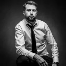 Czarno-biały portret brodatego mężczyzny w białej koszuli i krawacie, siedzącego na stołku z zegarkiem na nadgarstku, na ciemnym tle.