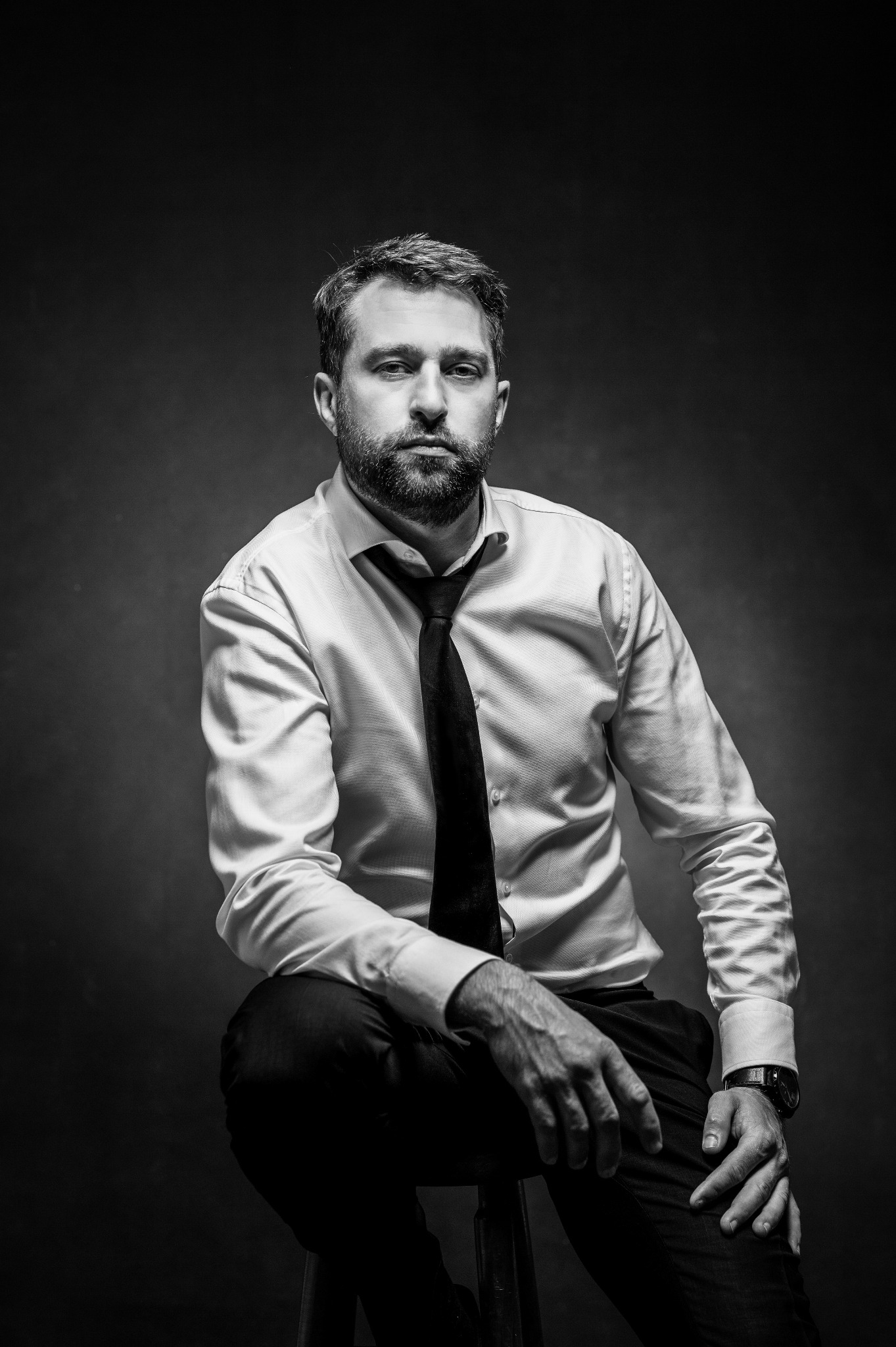 Czarno-biały portret brodatego mężczyzny w białej koszuli i krawacie, siedzącego na stołku z zegarkiem na nadgarstku, na ciemnym tle.