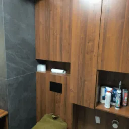 Drewniane szafki łazienkowe z ukrytą spłuczką i półkami na akcesoria toaletowe, na tle szarej ściany z płytek. Widoczny fragment toalety z ręcznikiem.