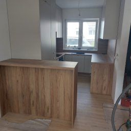 Meb-daw - Nowoczesna kuchnia z drewnianym blatem i białymi szafkami, widok z perspektywy baru kuchennego. Widoczny odkurzacz i osoba sprzątająca po montażu.