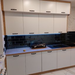 Meb-daw - Biała zabudowa kuchenna z czarnymi uchwytami i lustrzanymi płytkami nad blatem, podświetlona listwą LED. Uwagę zwraca wykończenie w kolorze drewna.