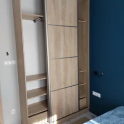 Szafa wnękowa z przesuwnymi drzwiami o strukturze drewna, z widocznym wnętrzem i półkami, na tle ściany w kolorze głębokiego granatu.