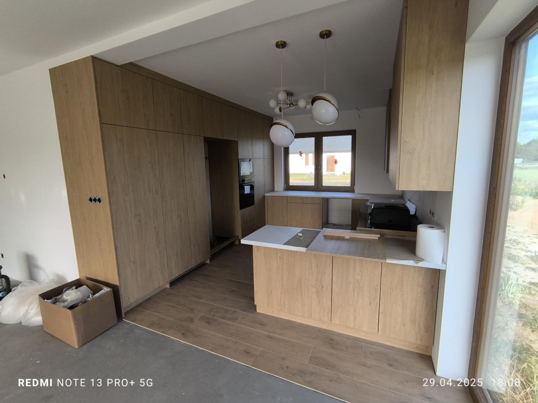 Jasna kuchnia z drewnianymi frontami w trakcie montażu, widoczne niedokończone podłogi i oświetlenie. Nowoczesny design w surowym wnętrzu.