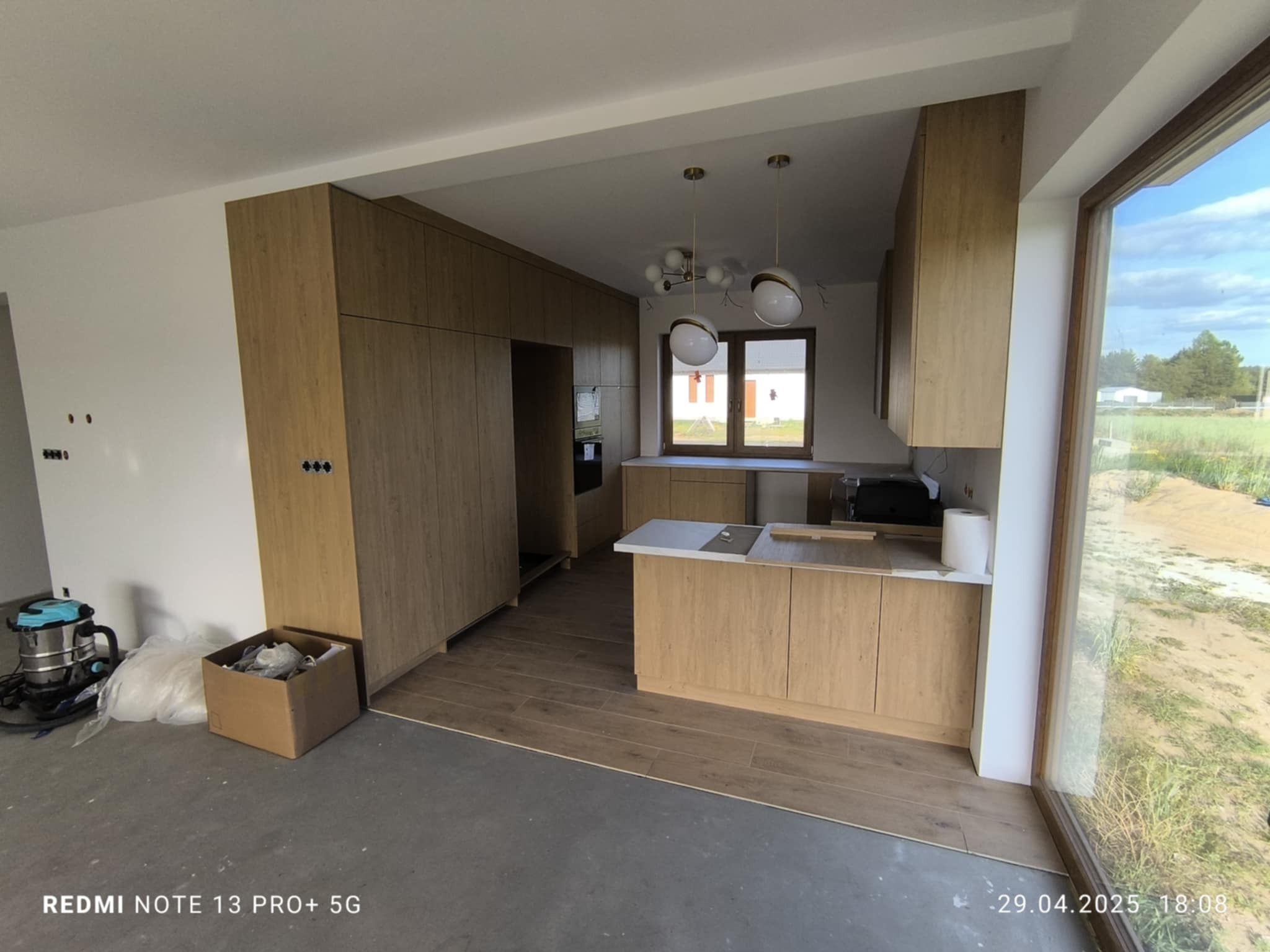 Jasna kuchnia z drewnianymi frontami, widok na zewnątrz przez duże okno. Podłoga częściowo wyłożona panelami. W tle nowoczesne lampy.