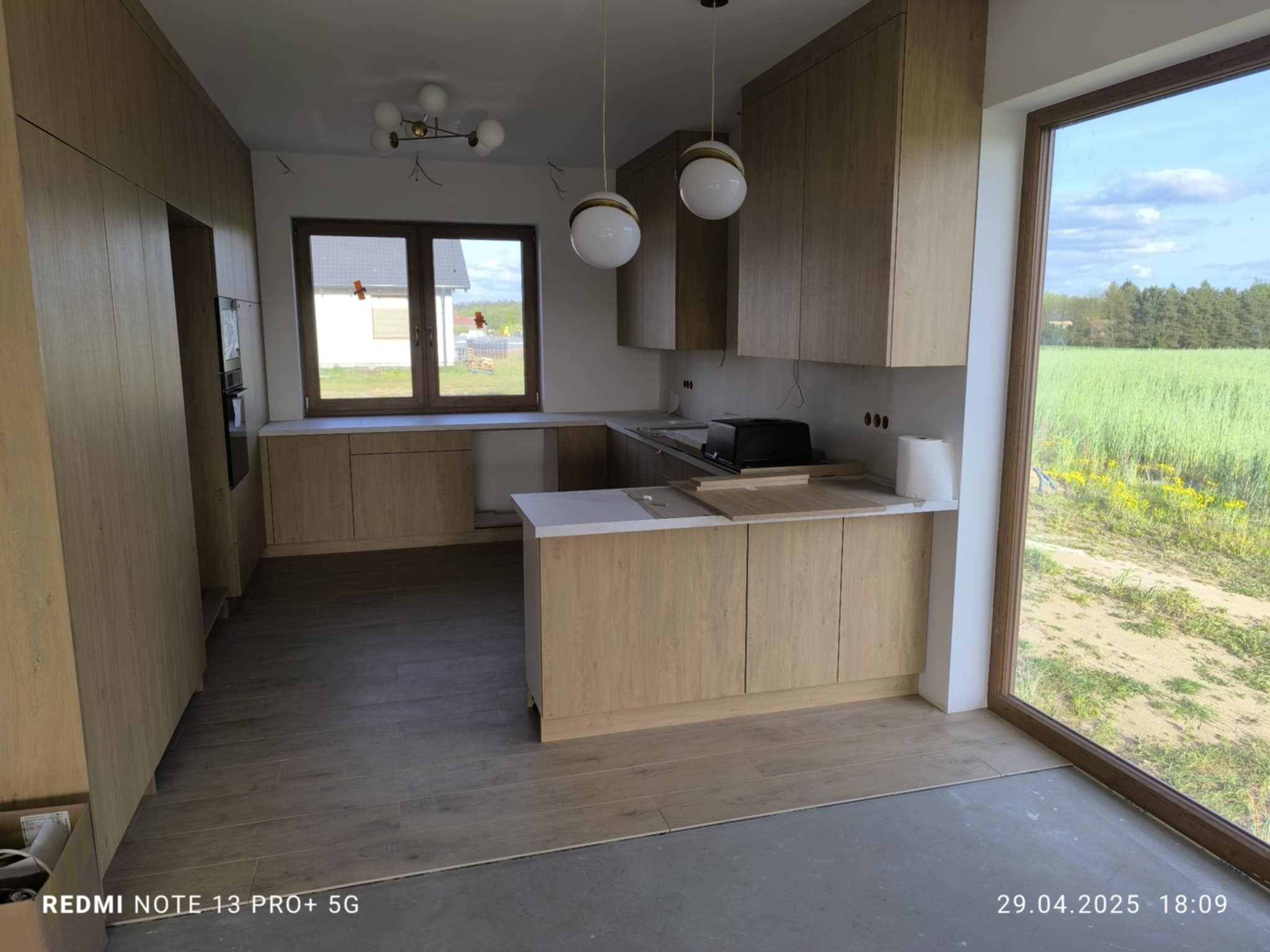 Jasna, nowoczesna kuchnia z drewnianymi frontami. Widoczne wiszące lampy kuliste, okno z widokiem na zewnątrz i częściowo ułożona podłoga z paneli.