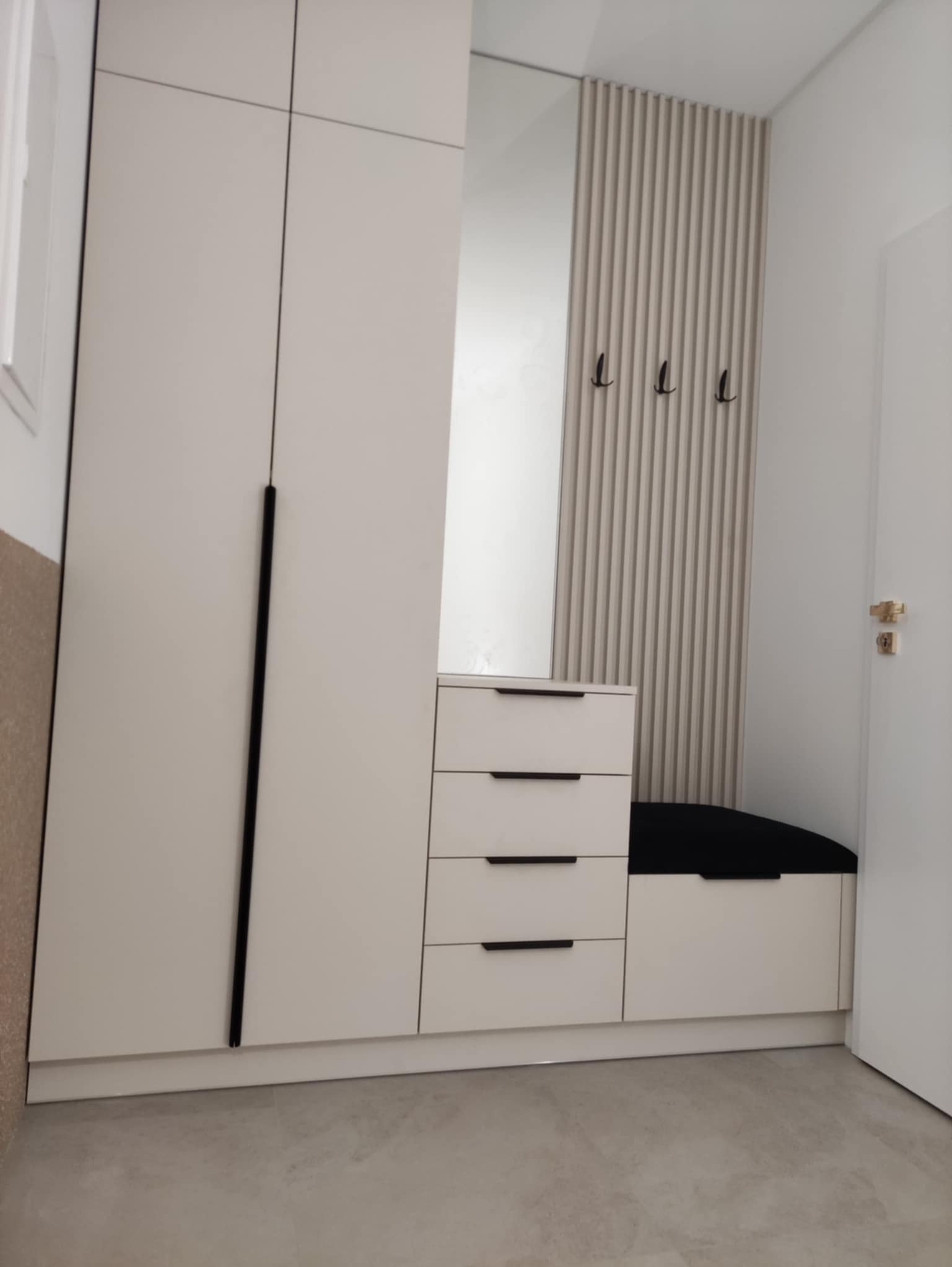 Jasna szafa wnękowa z lustrem, siedziskiem i wieszakami, minimalistyczny design. Pionowe lamele dekoracyjne. Czarne, długie uchwyty. Nowoczesne wykończenie przedpokoju.