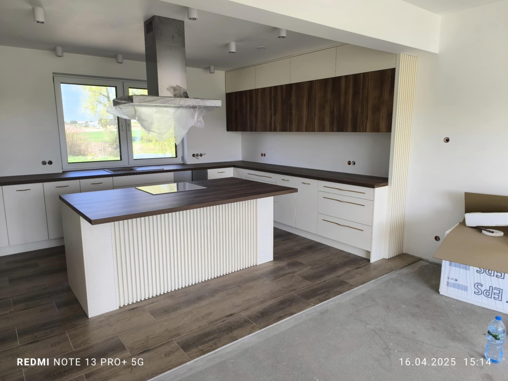 Nowoczesna kuchnia z wyspą w Kwilczu: białe fronty, drewniane akcenty, okap pod folią ochronną. Widoczne ułożone panele podłogowe i kartony po zabudowie.